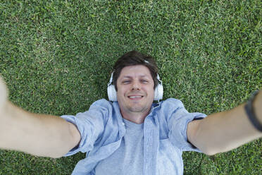 Happy man wearing headphones taking selfie and lying in grass - TYF00472