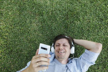 Happy man wearing headphones and using smart phone in grass - TYF00477