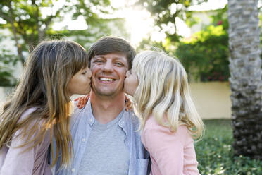 Happy father with daughters kissing him on cheeks at park - TYF00480