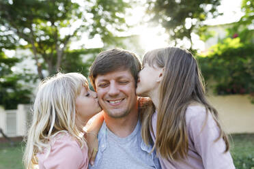 Smiling father with daughters kissing him on cheek at park - TYF00481