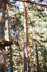 Kind in Sportkleidung auf einer Seilrutsche inmitten von Bäumen bei einem Wochenendtag im Abenteuerpark im Nadelwald - ADSF41473