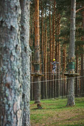 Kind, das auf einer Plattform auf einem Nadelbaum steht, während es einen Hindernisparcours in einem Abenteuerpark in einem sonnenbeschienenen Waldgebiet erkundet - ADSF41475