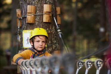 Junge mit gelbem Helm steht auf einer Hängebrücke in der Nähe eines Baumstamms an einem sommerlichen Wochenendtag im Abenteuerpark im Wald - ADSF41476