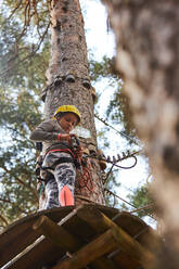 From below girl in activewear and helmet adjusting and examining belay while standing on round platform near coniferous tree on weekend day in adventure park - ADSF41477