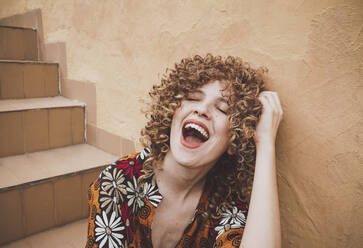 Fröhliche junge Frau mit lockigem Haar und roten Lippen sitzt mit geschlossenen Augen lächelnd im Treppenhaus an der Wand - ADSF41481