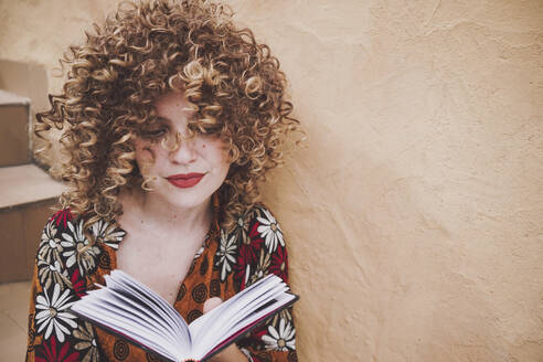 Junge Frau mit lockigem Haar mit aufgeschlagenem Buch und an die Wand gelehnt - ADSF41482