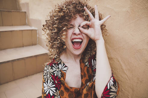 Erfreute junge Frau mit lockigem Haar zeigt okay Geste in der Nähe von Auge gegen Wand - ADSF41483