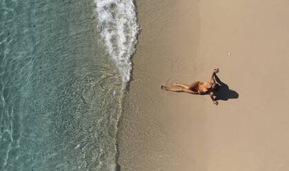 Luftaufnahme einer schönen Frau, die einen orangefarbenen Bikini trägt, während sie ein Sonnenbad am Sandstrand vor einem kristallklaren Meer in San Sostene, Kalabrien, Italien, nimmt. - AAEF16789