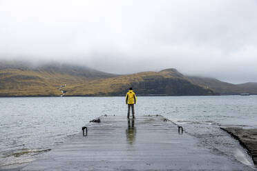 Man standing on pier in front of sea and mountains under cloudy sky - WPEF06817