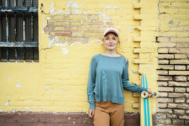 Lächelnde Frau mit Mütze steht mit Skateboard vor einer Wand - RCPF01530