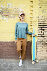 Thoughtful woman wearing cap leaning with skateboard on wall - RCPF01531