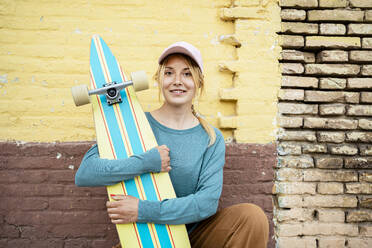 Smiling woman wearing cap crouching with skateboard in front of wall - RCPF01532