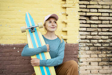 Thoughtful woman wearing cap crouching with skateboard in front of wall - RCPF01533