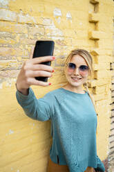 Lächelnde Frau, die ein Selfie durch ein Smartphone vor einer gelben Wand macht - RCPF01537
