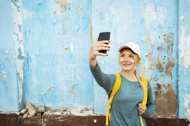 Lächelnde Frau mit Rucksack nimmt Selfie durch Smartphone vor der Wand - RCPF01545