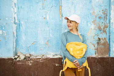 Nachdenkliche junge Frau mit gelbem Rucksack lehnt an blauer rissiger Wand - RCPF01546