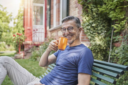 Happy man holding coffee cup on bench in garden - RORF03119