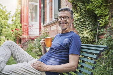 Glücklicher Mann mit Kaffeetasse auf einer Bank im Hinterhof sitzend - RORF03120