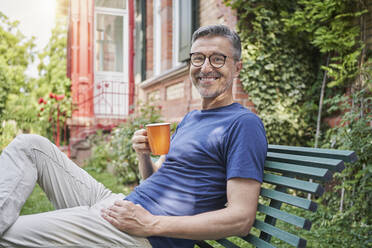 Happy man with coffee cup sitting on bench in back yard - RORF03120