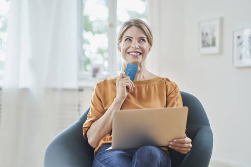 Nachdenklich lächelnde Frau mit Kreditkarte und Laptop auf einem Sessel zu Hause - RORF03123
