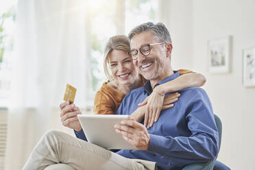 Smiling woman embracing man with credit card doing online shopping through tablet PC at home - RORF03134