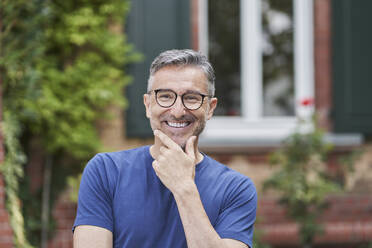 Smiling man wearing eyeglasses with hand on chin in front of house - RORF03141