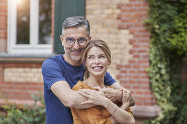 Smiling mature man embracing woman from behind in front of house - RORF03151