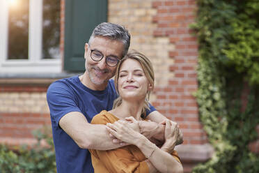 Smiling man embracing woman with eyes closed in front of house - RORF03152