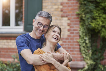 Smiling man embracing happy woman in front of house - RORF03153