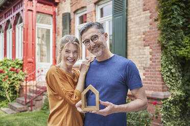 Smiling woman standing by man holding house model in garden - RORF03156
