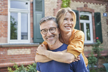 Happy woman embracing man from behind in front of house - RORF03161