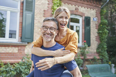 Smiling man giving piggyback ride to happy woman in garden - RORF03164