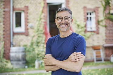 Glücklicher Mann mit Brille und verschränkten Armen im Hinterhof - RORF03172