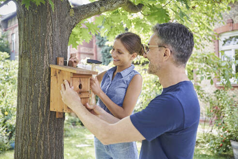 Lächelnder Mann mit Tochter hängt Nistkasten an Baumstamm im Garten auf - RORF03174
