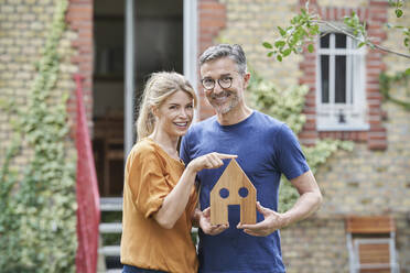 Happy man holding wooden house model by woman in garden - RORF03190