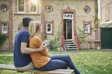Mann umarmt Frau mit Blick auf Haus auf Bank im Garten sitzend - RORF03194