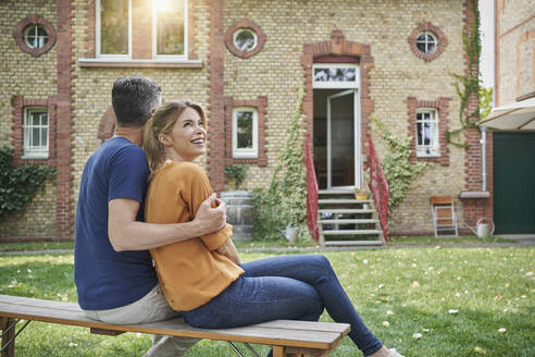 Mann umarmt glückliche Frau, die auf einer Bank im Hinterhof sitzt - RORF03195
