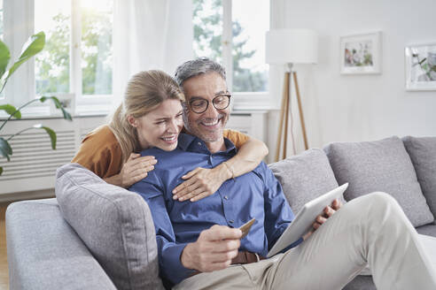 Frau mit Arm um Mann, der eine Kreditkarte hält, beim Online-Shopping über einen Tisch-PC auf dem Sofa - RORF03206