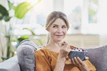Smiling woman holding yogurt bowl with fruits on sofa at home - RORF03207