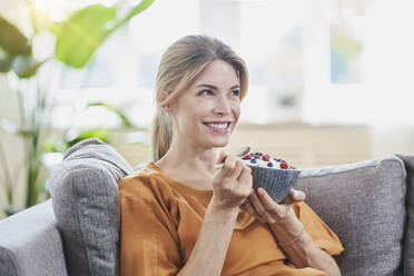 Happy woman holding yogurt bowl on sofa at home - RORF03208