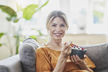 Happy woman holding yogurt bowl on sofa at home - RORF03209
