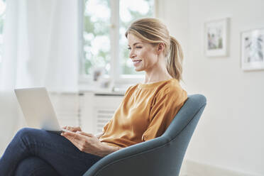 Smiling businesswoman using laptop in armchair at home - RORF03228