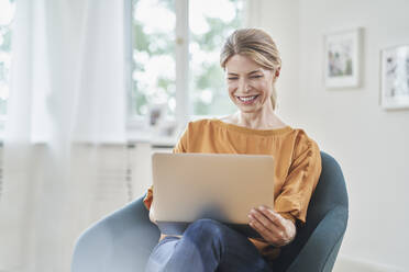 Happy businesswoman using laptop in armchair at home - RORF03230