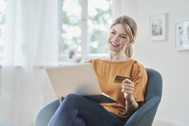 Smiling woman holding credit card doing online shopping through laptop in armchair at home - RORF03233