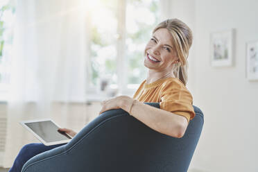 Lächelnde Frau mit Tablet-PC im Sessel zu Hause - RORF03236