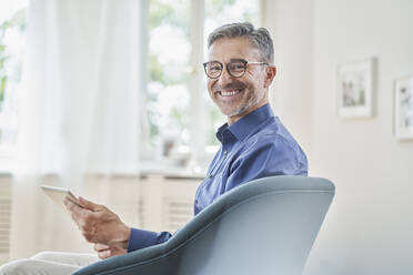 Happy businessman holding tablet PC in armchair at home - RORF03237