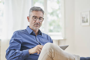 Mature businessman looking at tablet PC in armchair at home - RORF03238