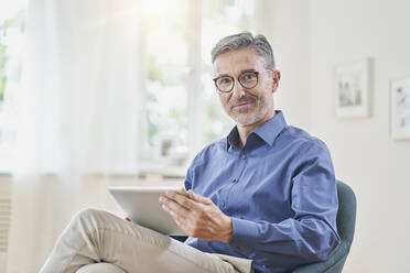 Smiling businessman holding tablet PC in armchair at home - RORF03239