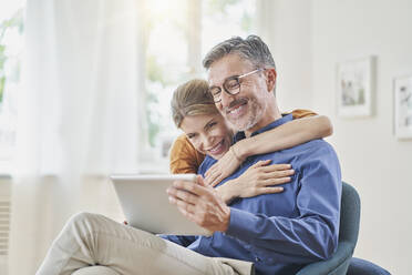 Ein glücklicher Mann zeigt einer Frau im Sessel zu Hause einen Tablet-PC - RORF03241
