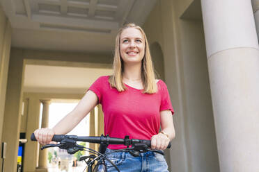 Glückliche Frau steht mit Fahrrad vor einem Gebäude - TAMF03631